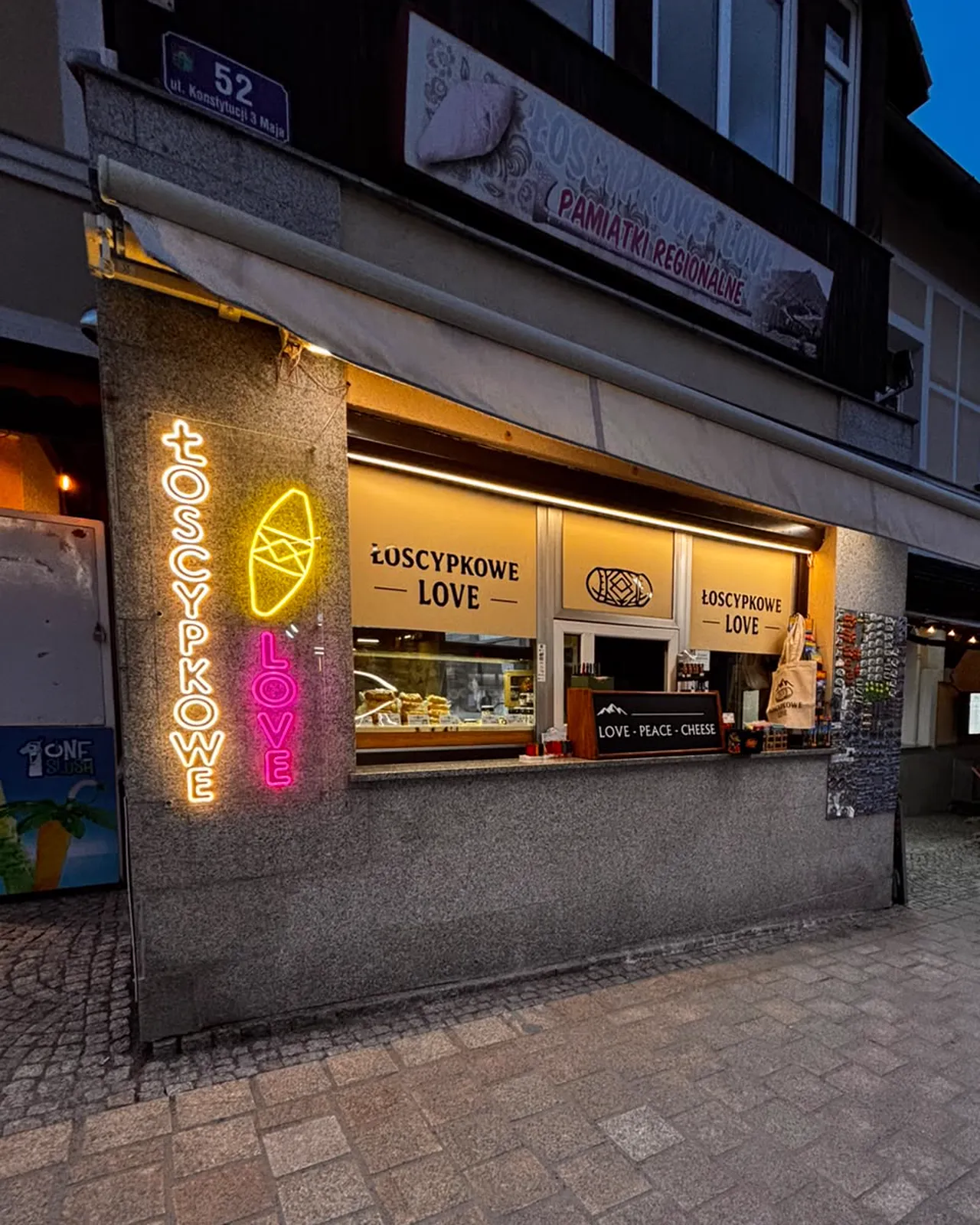 Róg sklepu z dwoma neonami i numerem 52, obok okienko z serkami i markiza