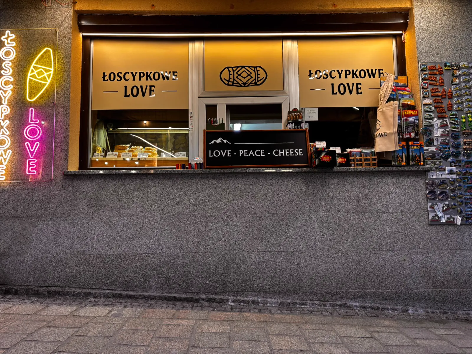 Okienko sklepu Łoscypkowe Love wieczorem: serki w podświetlonej ladzie, tabliczka Love Peace Cheese, po lewej neon, po prawej magnesy